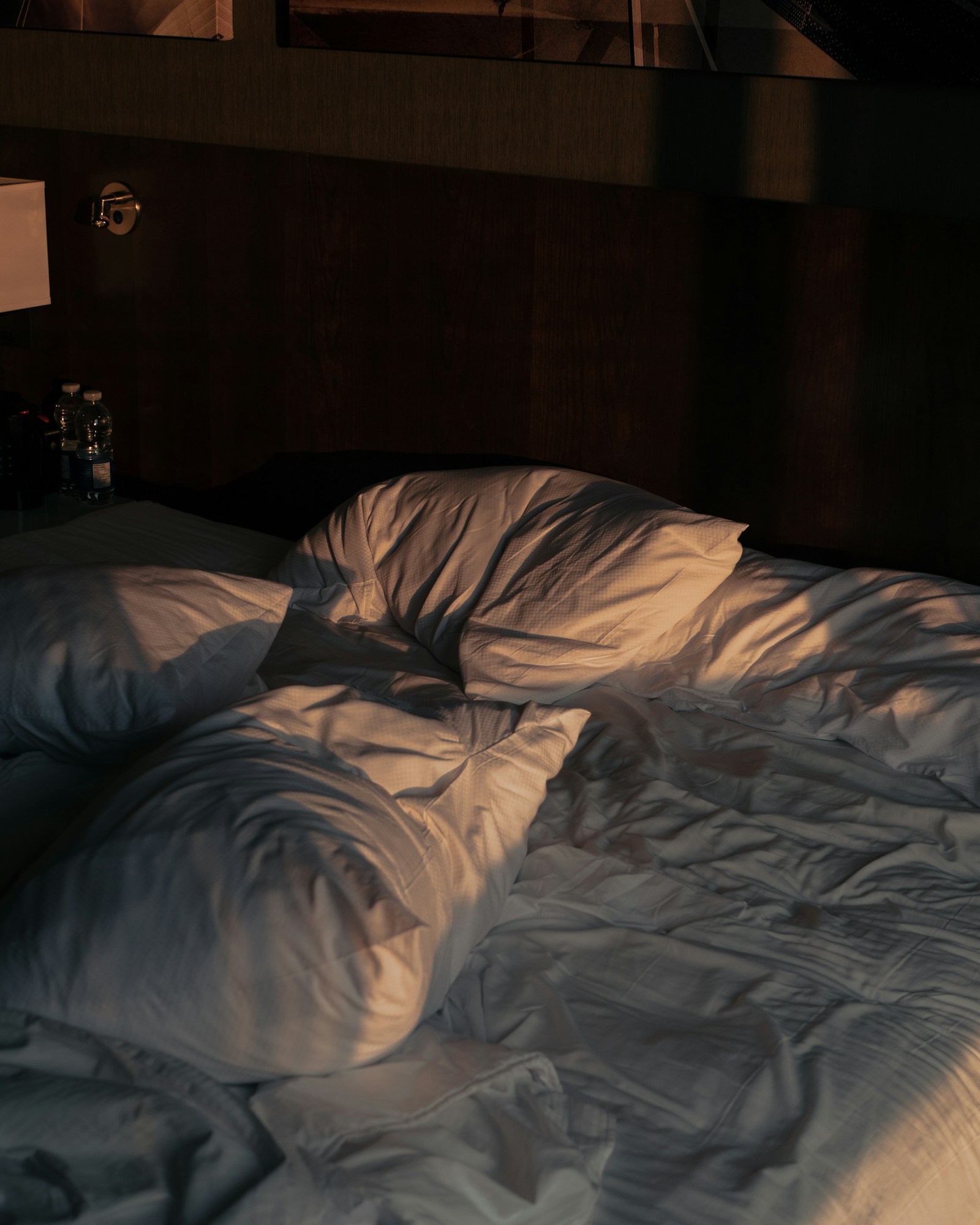 Cama de hotel desfeita com lençóis brancos amarrotados e cabeceira de madeira escura ao fundo — costuras, ripas e cantos do estrado.