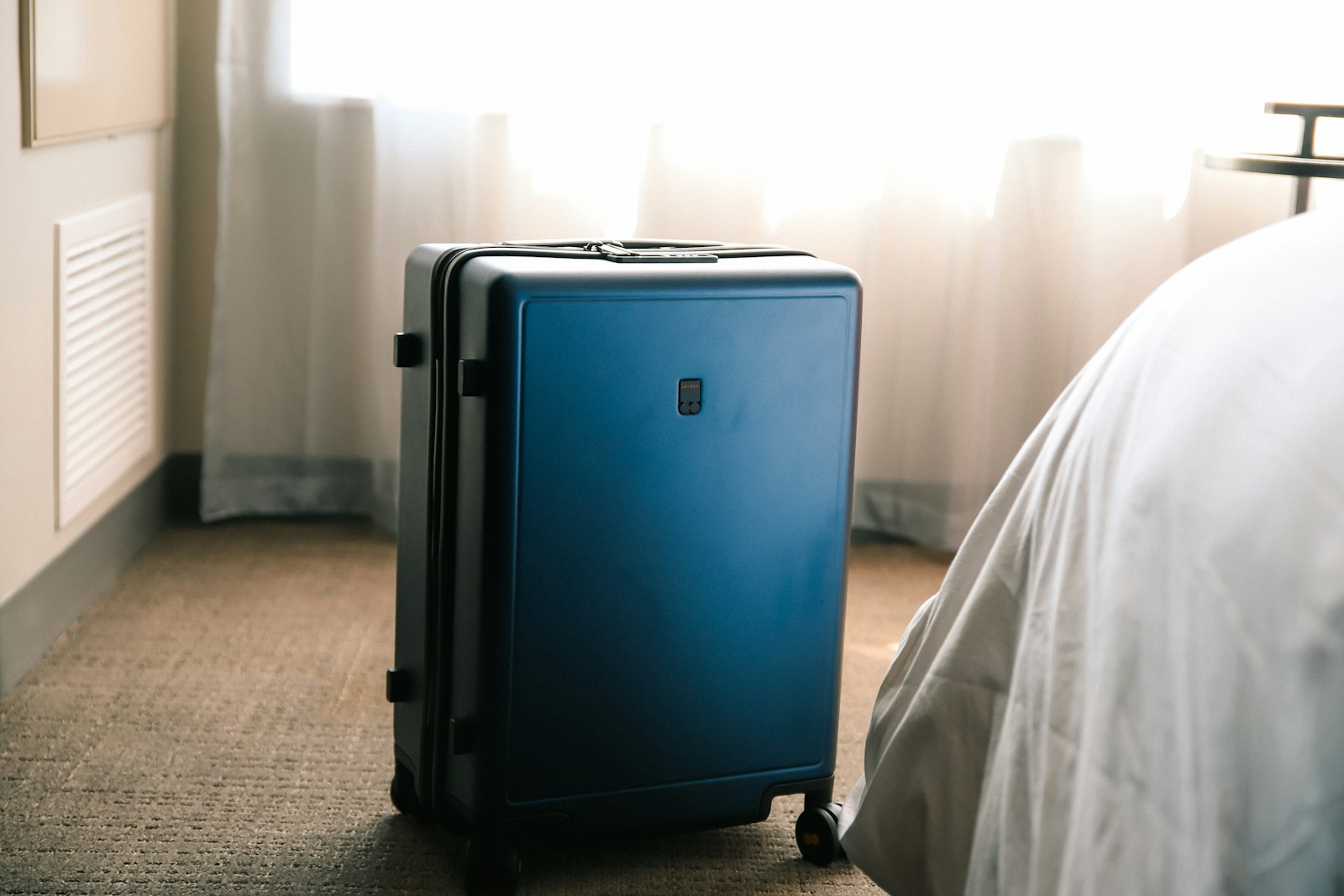 Mala azul de viagem em pé junto à cama de hotel com lençóis brancos — o risco de transporte de percevejos entre quarto e casa.