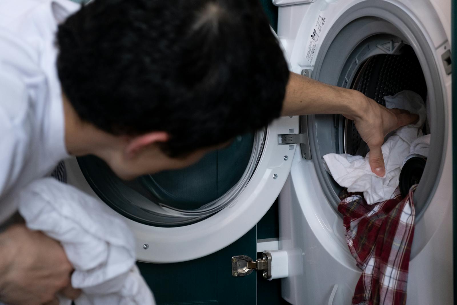 Pessoa a colocar roupa de cama branca no tambor de uma máquina de lavar — primeiro passo antes de inspecionar e aplicar vapor na estrutura.